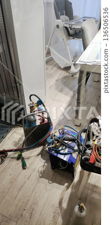 Refrigerator back panel removed, compressor and condenser coils exposed for repair. Refrigeration tools, gas cylinders, manifold gauge set, and vacuum pump rest on wood floor. 136506536