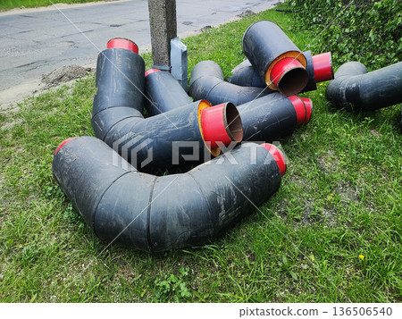 Insulated black pipes red interior material on green grass, construction equipment beside damaged asphalt road concrete pole infrastructure project 136506540