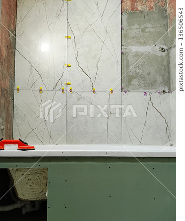 Bathroom renovation with new ceramic wall tiles installation above white bathtub; construction tools visible for tiling work; unfinished wall surface; plumbing elements below tub 136506543