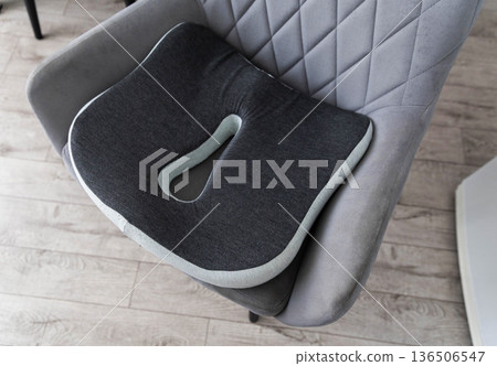 Modern ergonomic orthopedic seat cushion, dark grey color, light grey border, positioned on grey upholstered chair indoors. Modern ergonomic orthopedic seat cushion, dark grey color, light grey border, positioned on grey upholstered chair indoors. 136506547