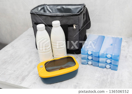 Cooler bag with two bottles of liquid, multiple ice packs, and yellow lunchbox on light surface preparing for transport of cold items 136506552