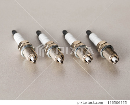 Four automotive spark plugs arranged diagonally on muted gray surface, presenting metallic threading and ceramic insulator for engine ignition systems. Four automotive spark plugs arranged diagonally on muted gray surface, presenting metallic threading and ceramic insulator for engine ignition systems. 136506555