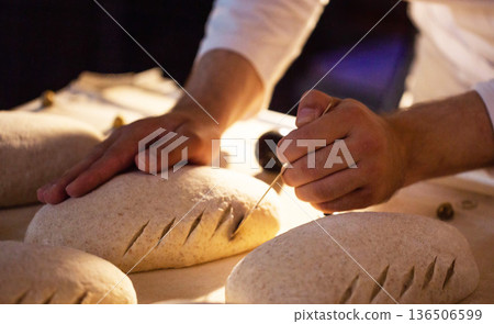 Manual scoring of unbaked bread dough loaves by baker hand in artisanal bread factory. Traditional preparation of raw bread for baking. Manual scoring of unbaked bread dough loaves by baker hand in artisanal bread factory. Traditional preparation of raw bread for baking. 136506599