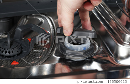Close up view of human hand unscrewing and lifting black cap from salt reservoir in bottom of modern dishwasher appliance Close up view of human hand unscrewing and lifting black cap from salt reservoir in bottom of modern dishwasher appliance 136506613