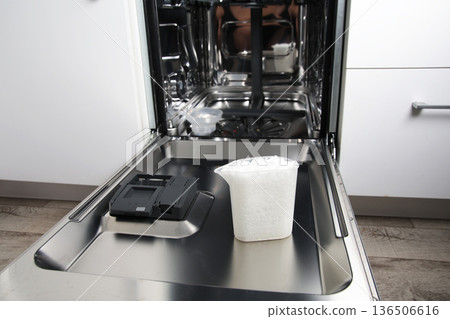 Open dishwasher interior, clean stainless steel. Measuring cup with white granular salt on lower door. Essential water softening agent for kitchen appliance 136506616
