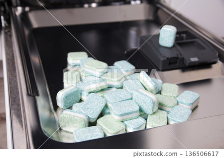 Many dishwasher tablets, green and blue, piled on door of open dishwasher machine, illustrating cleaning detergent for dishwashing appliance. 136506617