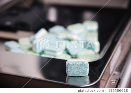 Close up view of dishwasher detergent tablet resting on rack of kitchen appliance, background with many cleaning pods blurred 136506618