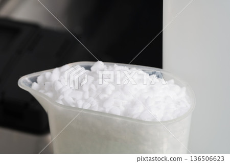White granular salt in plastic measuring cup for dishwasher, showing importance of specialty salt for optimal machine performance and sparkling dishes. 136506623