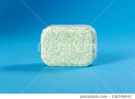 Single green dishwasher tablet in transparent plastic packaging against solid blue background ready for household cleaning use. 136506641