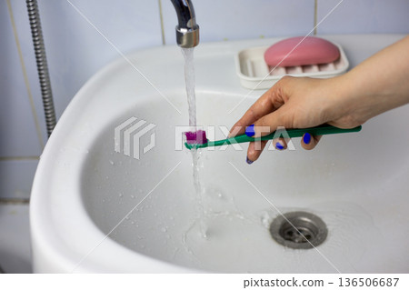 Toothbrush under running water. Hand holds brightly colored toothbrush under sink faucet. Stream of water washes away dirt from toothbrush after brushing. Lived-in environment 136506687