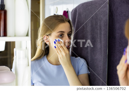Young European woman uses cotton pad soaked in makeup remover in front of bathroom mirror. Process of removing makeup after day. Skincare routine for removing makeup in front of bathroom mirror Young European woman uses cotton pad soaked in makeup remover in front of bathroom mirror. Process of removing makeup after day. Skincare routine for removing makeup in front of bathroom mirror 136506702