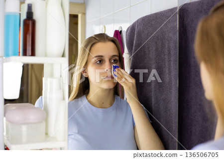 Young woman removes makeup with cotton pad in front of mirror. Micellar water on pad removes eyeshadow. Young woman in cosmetic routine removes makeup from her face, caring for her skin Young woman removes makeup with cotton pad in front of mirror. Micellar water on pad removes eyeshadow. Young woman in cosmetic routine removes makeup from her face, caring for her skin 136506705