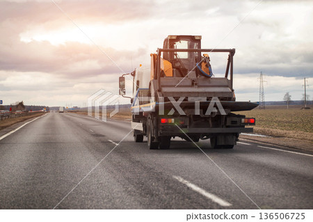 Tow truck transporting skid steer loader on highway under cloudy sky, emphasizing logistics of heavy equipment delivery and industrial transport needs. Copy space for text 136506725