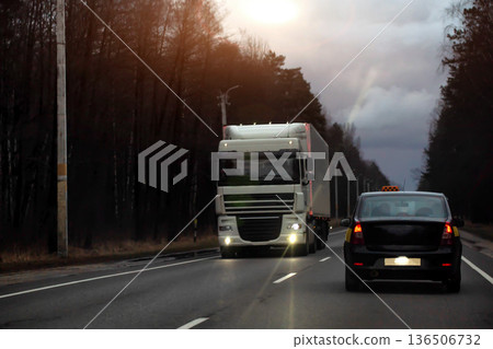 Nighttime road scene with semi-trailer truck driving towards camera on highway while black taxi car travels ahead, showing logistics and freight delivery. Copy space for text 136506732