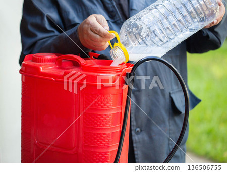 Person pouring liquid preparation from plastic bottle into red garden sprayer tank for plant treatment against pests agriculture horticulture farming 136506755