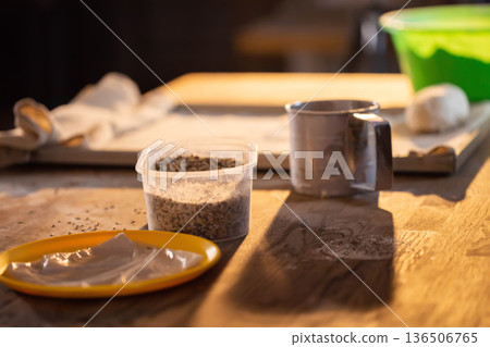 Kitchen table scene shows food preparation ingredients with granular seeds in plastic container, measuring cup, and yellow plate holding light-colored dough or paste, featuring bright soft light Kitchen table scene shows food preparation ingredients with granular seeds in plastic container, measuring cup, and yellow plate holding light-colored dough or paste, featuring bright soft light 136506765