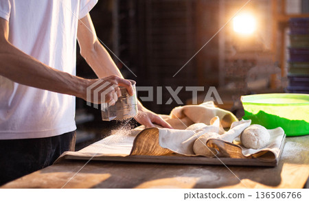 Person sifting flour onto dough portions arranged on linen cloth in tray, preparing artisan bread for baking process, warm sunlight illuminating kitchen or bakery setting, close up of hands and baking Person sifting flour onto dough portions arranged on linen cloth in tray, preparing artisan bread for baking process, warm sunlight illuminating kitchen or bakery setting, close up of hands and baking 136506766