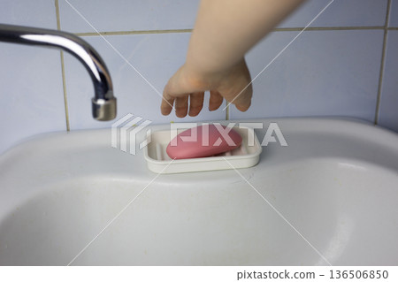 Girl hand reaches for pink soap in white soap dish. Concept of hand washing and skin hygiene. Sink in lived in room with less than ideal living conditions. Soap on sink 136506850