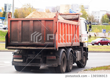Rear view of heavy dump truck with cargo bed full of sand on urban road, construction vehicle transporting material, autumn trees and buildings in background 136506913
