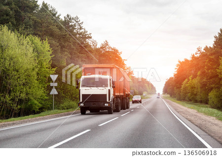Large white and orange truck driving on highway, flanked by green forest. Represents freight transport, logistics, industry, and movement of goods under cloudy sky. 136506918