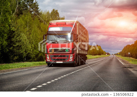Red cargo truck with trailer driving on asphalt highway through green forest landscape under cloudy sky in daytime, transportation 136506922