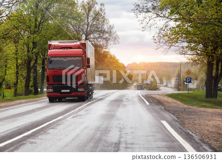 Red semi-truck travels on wet asphalt road lined with lush green trees on a cloudy spring day with a tractor parked roadside 136506931