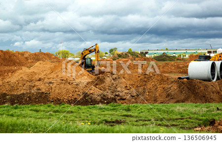 Yellow excavator moving earth for road construction. Infrastructure development project with soil piles and concrete pipes under cloudy sky. Yellow excavator moving earth for road construction. Infrastructure development project with soil piles and concrete pipes under cloudy sky. 136506945