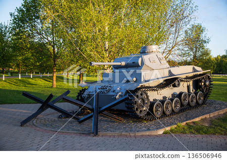 Historical military tank monument displayed in a public park setting during golden hour, featuring a grey battle tank and anti-tank obstacles surrounded by green trees and grass 136506946