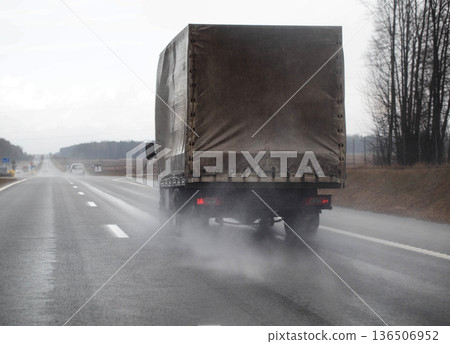 Large cargo truck driving away on wet highway during cloudy day, water spray trailing from rear wheels, long transport vehicle moving on open road surface. Copy space for text Large cargo truck driving away on wet highway during cloudy day, water spray trailing from rear wheels, long transport vehicle moving on open road surface. Copy space for text 136506952
