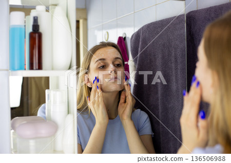 Girl rubs facial cleansing foam into her hands. Young woman with blonde hair performs her beauty routine and skin care in front of bathroom mirror. Maintaining healthy facial skin Girl rubs facial cleansing foam into her hands. Young woman with blonde hair performs her beauty routine and skin care in front of bathroom mirror. Maintaining healthy facial skin 136506988