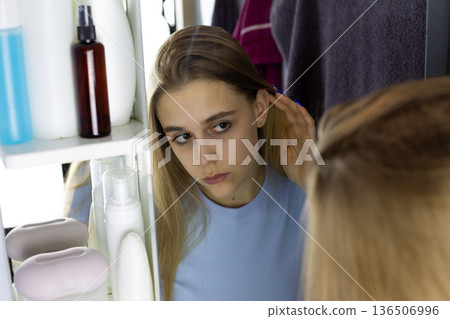 Young girl looks at her face in bathroom mirror with interest. Frustrated girl looks at her facial skin. Skin care and beauty concept for woman. Girl morning routine in mirror 136506996