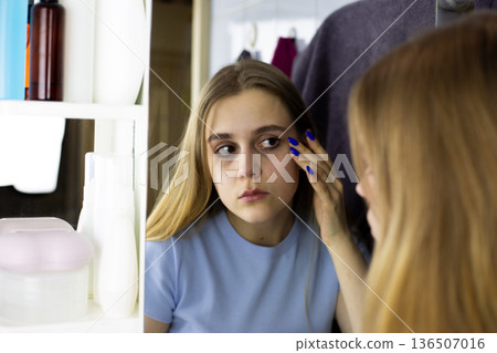 Natural girl looking on her skin. Realistic young blonde carefully examines condition of her skin. Facial hygiene concept. Girl looks at herself in mirror reflection in bathroom Natural girl looking on her skin. Realistic young blonde carefully examines condition of her skin. Facial hygiene concept. Girl looks at herself in mirror reflection in bathroom 136507016