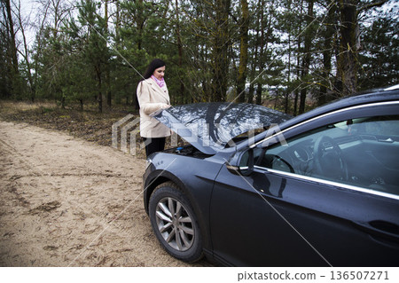 Woman stands next to broken-down car with open hood on dirt road in pine forest, inspecting engine compartment, experiencing vehicle trouble outdoors during day Woman stands next to broken-down car with open hood on dirt road in pine forest, inspecting engine compartment, experiencing vehicle trouble outdoors during day 136507271