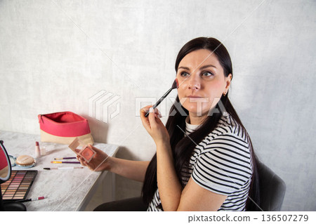Woman applies facial makeup blush using brush at home vanity table, dark hair person beauty routine indoors, cosmetic application process. Copy space for text Woman applies facial makeup blush using brush at home vanity table, dark hair person beauty routine indoors, cosmetic application process. Copy space for text 136507279