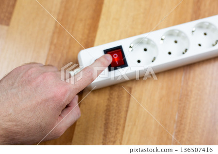 Person hand is shown turning on white extension cord by flipping its red switch, which lies on rustic wooden plank. Simple, everyday actions in home or workshop environments Person hand is shown turning on white extension cord by flipping its red switch, which lies on rustic wooden plank. Simple, everyday actions in home or workshop environments 136507416