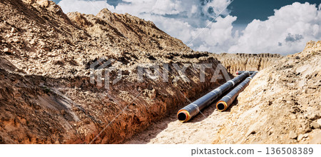 Workers are laying large pipes in a deep trench on a construction site in a rural area. The sky is blue with some clouds above 136508389