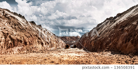 Workers are digging a trench at a construction site. Large pipes are placed at the bottom of the trench. Clouds cover the sky above the site 136508391