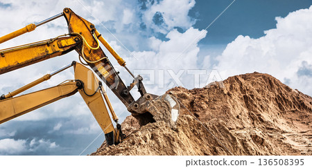 An excavator is digging into a large pile of dirt at a construction site. The sky has clouds, and it is daytime. Work is in progress An excavator is digging into a large pile of dirt at a construction site. The sky has clouds, and it is daytime. Work is in progress 136508395