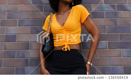 Young woman stands against a brick wall wearing a bright top and black pants while holding a bag Young woman stands against a brick wall wearing a bright top and black pants while holding a bag 136508419