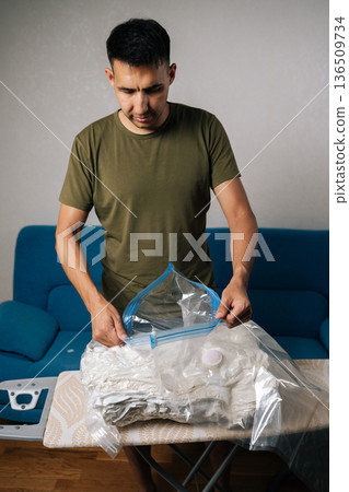 Man preparing to using vacuum cleaner to efficiently suck air out of space saver storage bag filled with bedding, maximizing storage space and promoting organization in home. 136509734