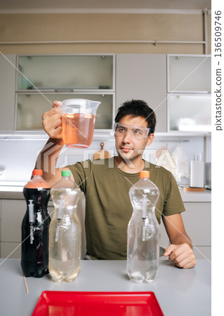 Vertical portrait of chemist in safety glasses holding beaker with liquid fertilizer, prepared from various chemicals in laboratory, emphasizing fertilizer preparation for agriculture and science. 136509746