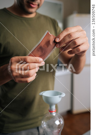 Vertical cropped shot of gardener pouring fertilizer from sachet into bottle using funnel, preparing nutrient solution for plants, demonstrating simple yet effective method for plant care at home. 136509748