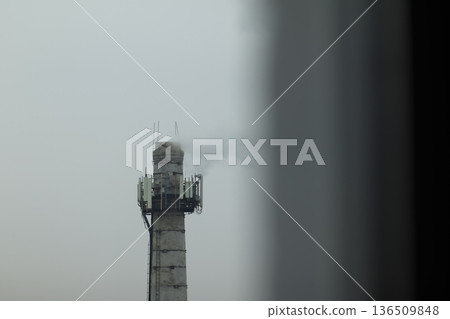 Industrial Chimney with Communication Antennas. Industrial concrete chimney with smoke and telecommunication antennas under a grey sky, urban pollution concept 136509848