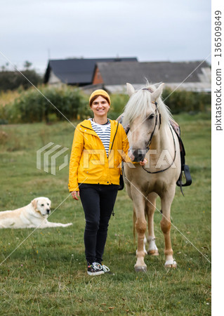 Country Life with Horse and Dog. A full-length portrait of a smiling woman in a yellow jacket and beanie standing next to a light-colored, palomino horse. Vertical 136509849