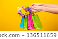 Hands holding colorful shopping bags filled with spring flowers against a vibrant yellow background 136511659