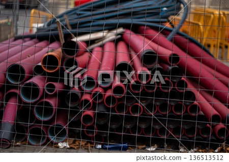 Stacked red corrugated pipes and blue spooled hoses behind a wire fence, awaiting installation for urban infrastructure development and utility construction work 136513512