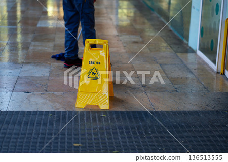 Yellow caution sign warning about a wet floor in a public building, a worker mopping the shiny tiled surface nearby, emphasizing cleaning, safety, and hygiene 136513555