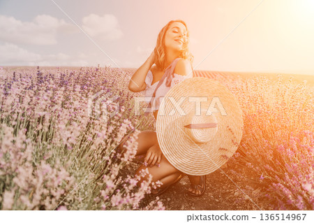 Lavender woman hat. Young woman enjoys the sunset in a blooming purple lavender field while holding a large straw hat Lavender woman hat. Young woman enjoys the sunset in a blooming purple lavender field while holding a large straw hat 136514967