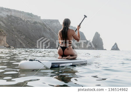 Woman Paddleboarding Ocean Coastline - A woman kneels on a paddleboard in the ocean, facing a rocky coastline. The sun is shining and the water is calm. 136514982