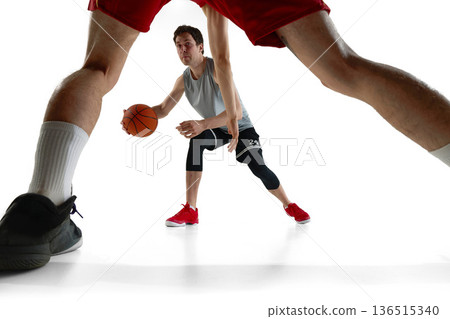 Low angle basketball player dribbling framed by defender legs isolated on white background. 136515340
